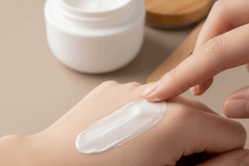 Applicazione di una crema cosmetica bianca su una zona arrossata della pelle del braccio, con vasetto di crema e spatola in legno su sfondo beige neutro