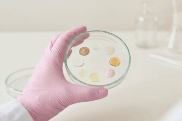 test sui cosmetici, cosmetici su piastra petri per analisi di laboratorio. Mano con guanto rosa in nitrile.